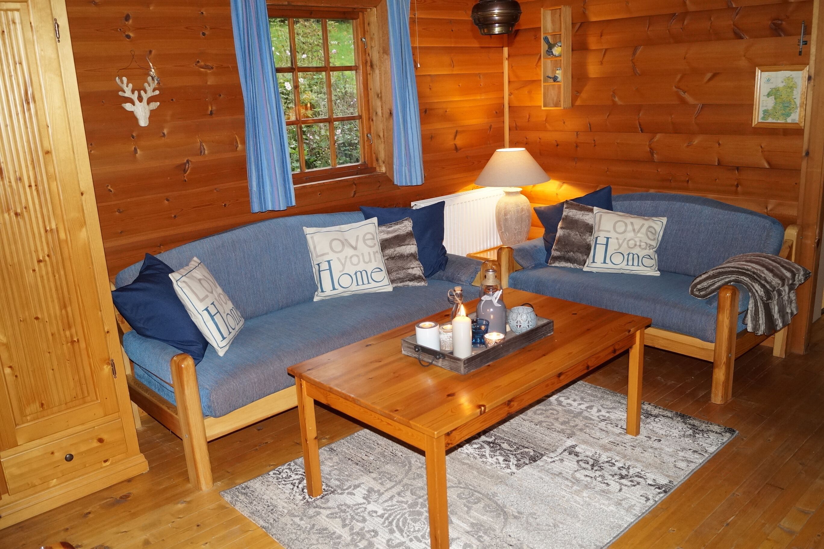 Un interior de cabaña acogedor con suelo de madera, un sofá azul con cojines que dicen 'Love Your Home', una mesa de centro de madera con velas y una ventana con cortinas azules.