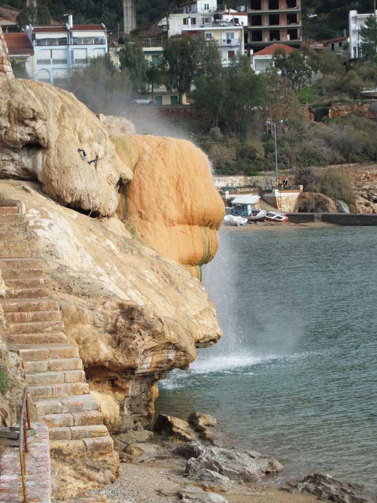 A natural rock formation with a water cascade, a staircase, and a view of buildings in the background.