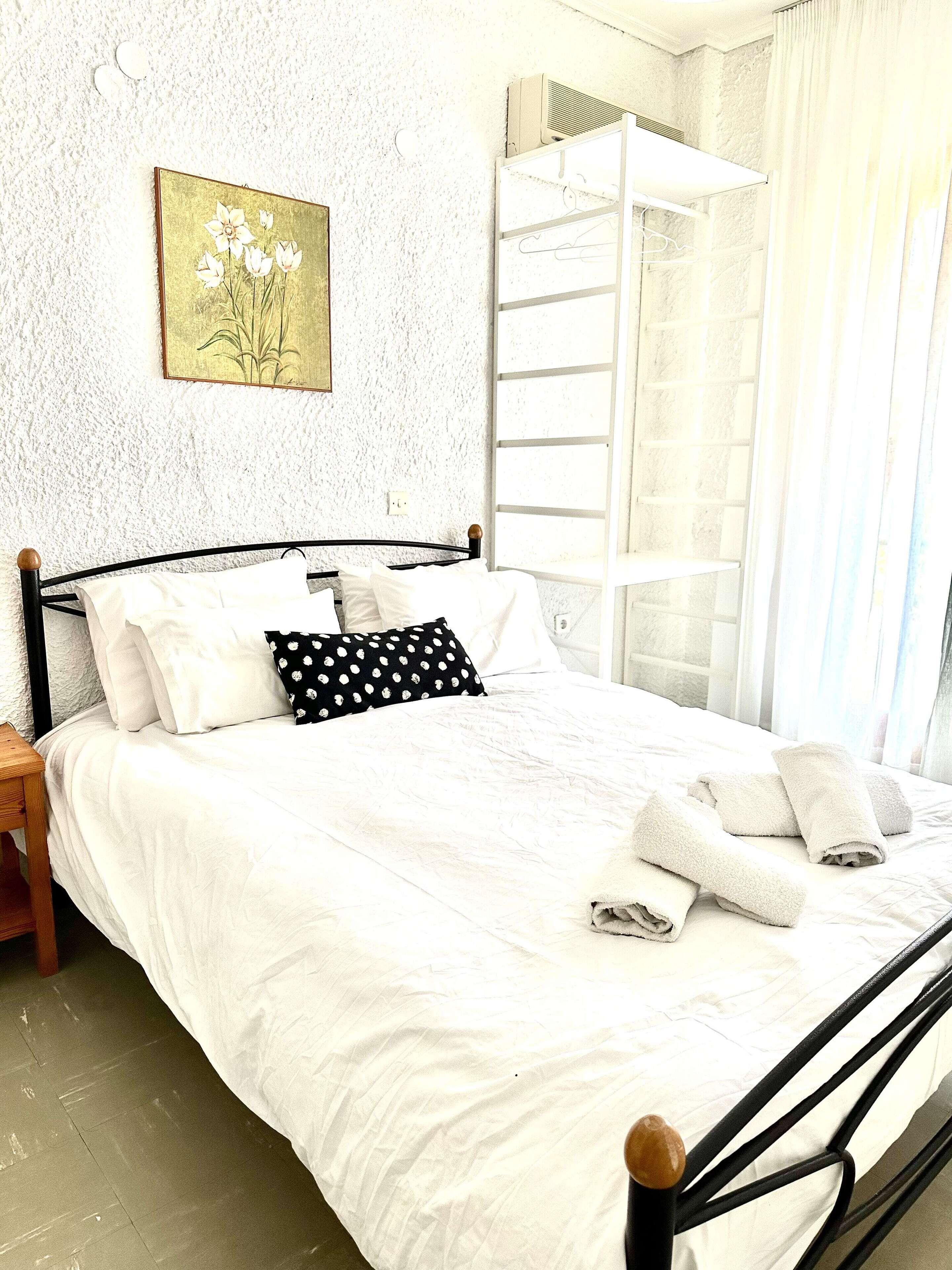 Een net opgemaakt bed met witte lakens en een zwart-wit stippenkussen, een houten nachtkastje, een wandgemonteerde airconditioning en een raam met een witte luxaflex.