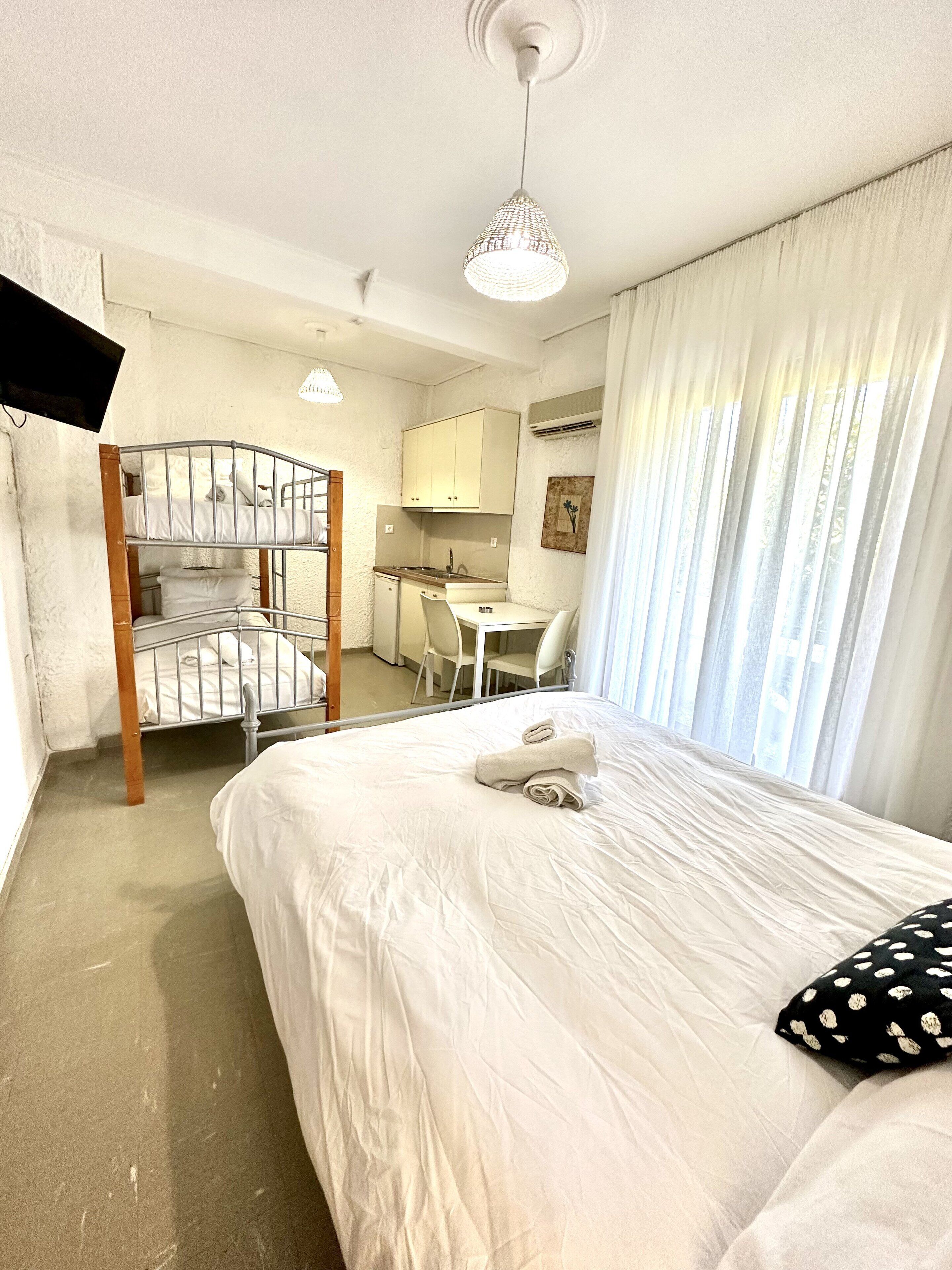 A compact hotel room with a bunk bed, a small kitchenette, a desk, and a bed with rolled towels.