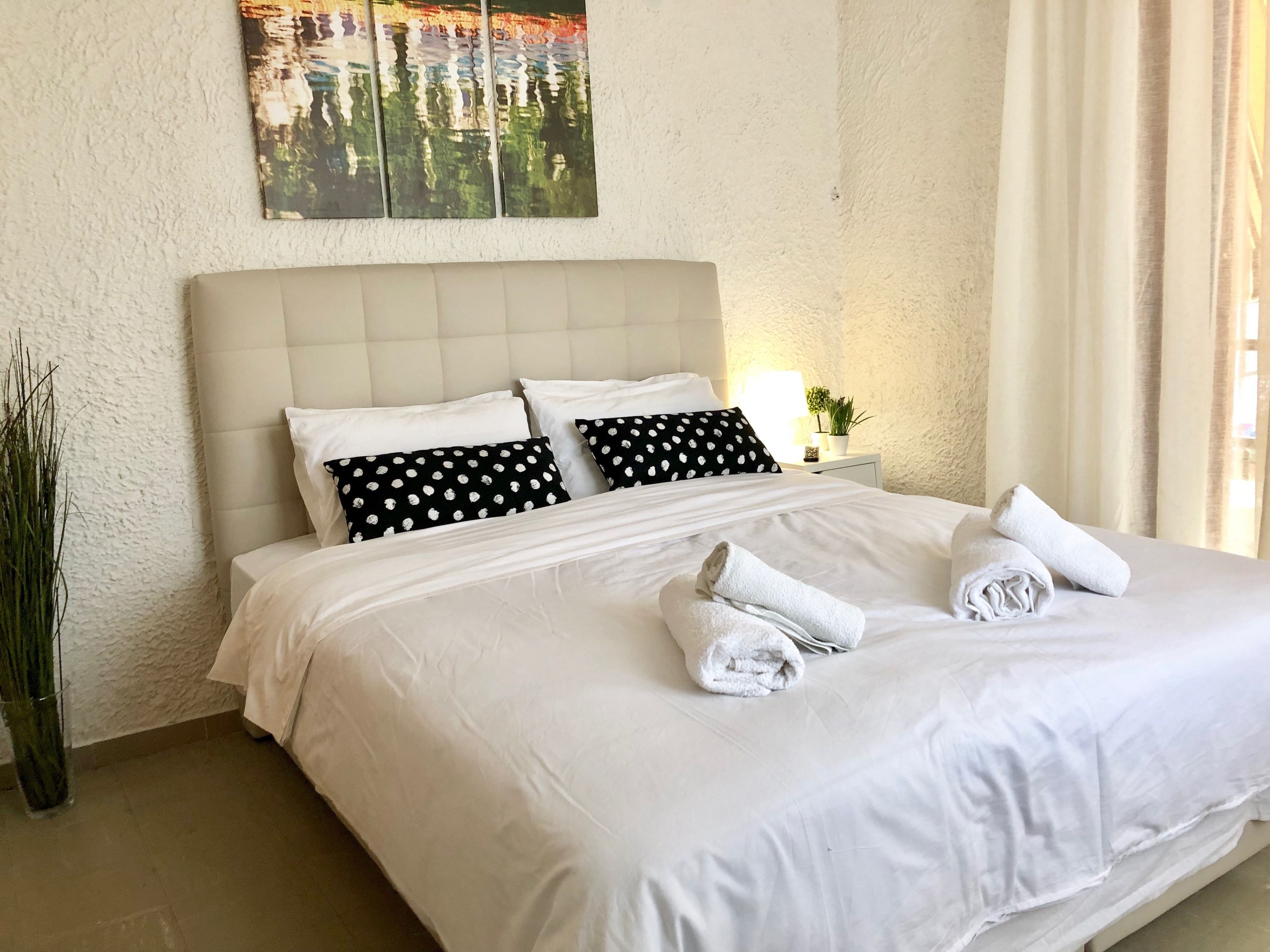 Ein ordentlich bezogenes Bett mit weißen Leinen und zwei schwarzen Kissen mit weißen Punkten, ein Nachttisch mit Lampe und einer kleinen Pflanze, sowie eine strukturierte Wand mit einem gerahmten Kunstwerk darüber.