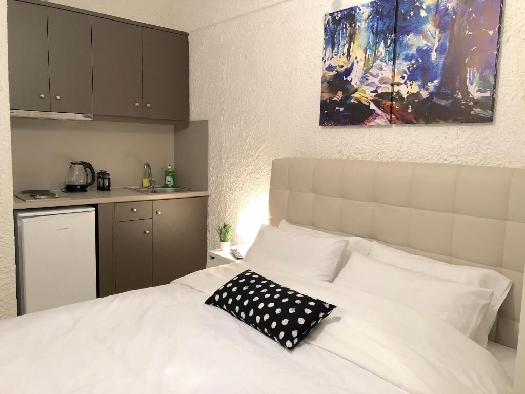 A bedroom with a bed, a nightstand, a painting on the wall, and a kitchenette with a refrigerator.