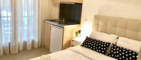A bedroom with a bed, pillows, a bedside table, a small kitchenette, and a window with curtains.