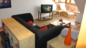 Un salon avec un canapé noir, une table basse en bois, un téléviseur et une fenêtre donnant sur l’extérieur.
