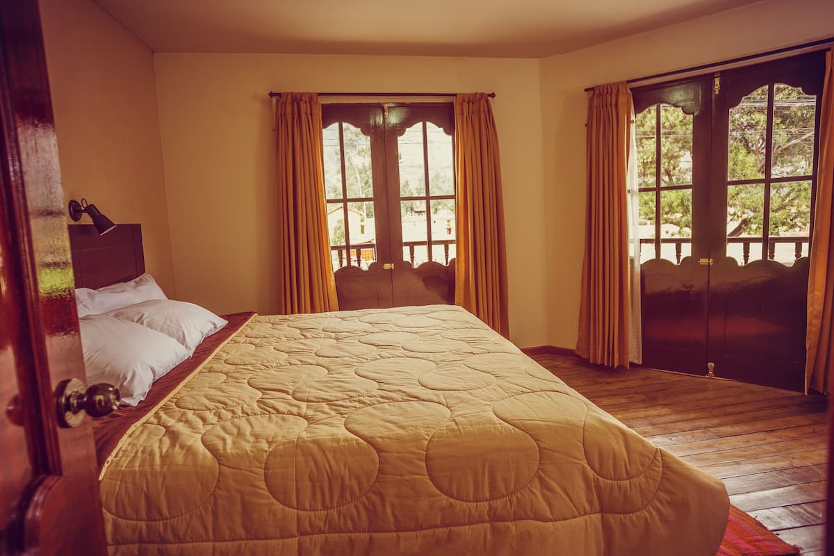 Ein Zimmer mit einem großen Bett, Holzboden und zwei mit Vorhängen versehenen Fenstern.