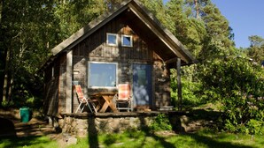 Una cabana rústica amb fonaments de pedra, façana de fusta i un porxo amb dues cadires.