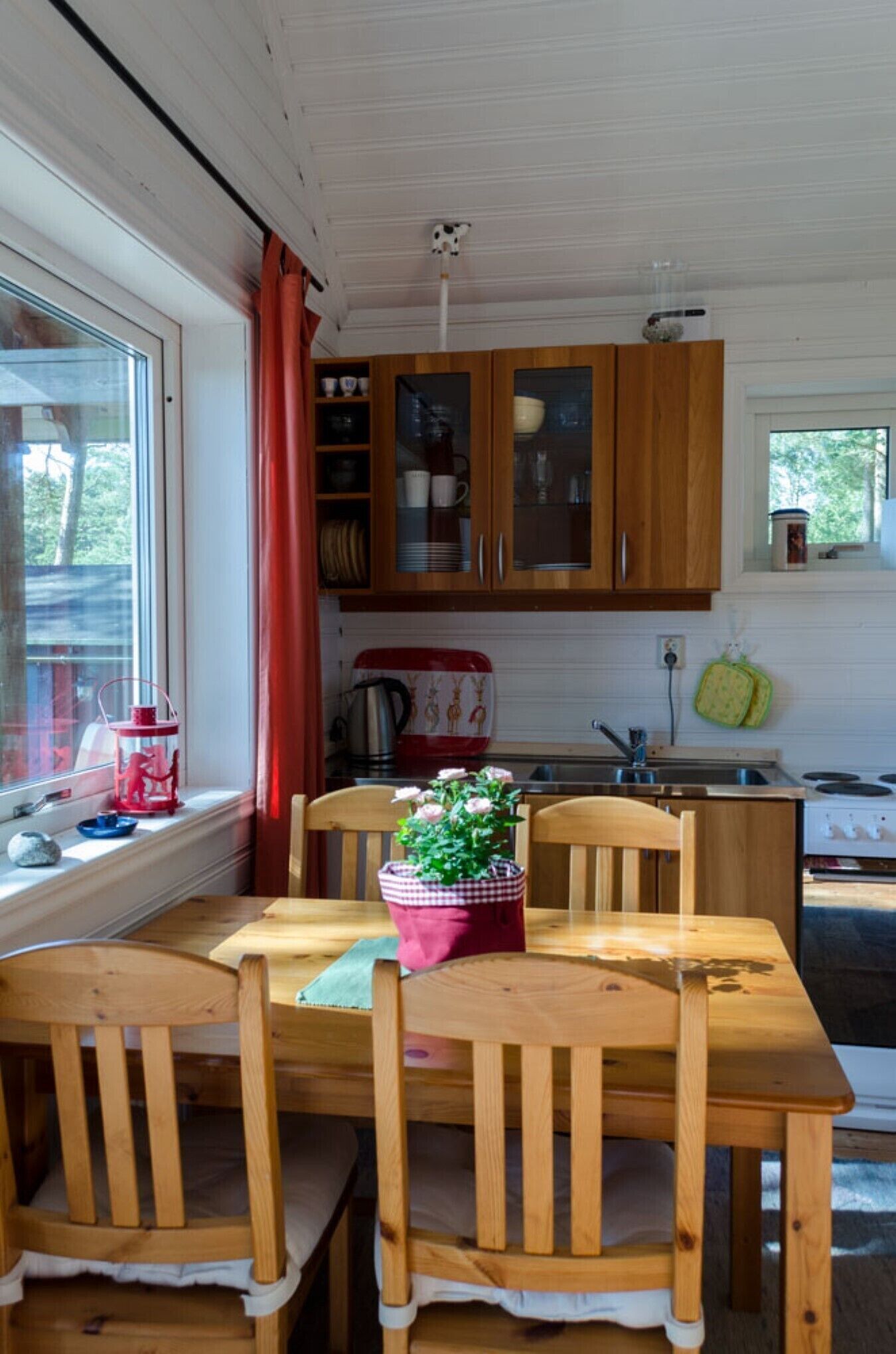 Una mesa de comedor de madera con sillas, una ventana con cortinas, armarios de madera y una zona de cocina con estufa y fregadero.