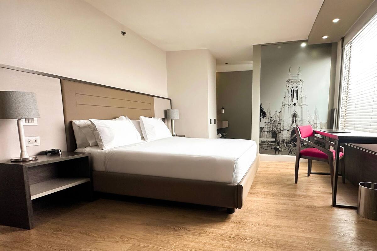 A hotel room with a bed, bedside lamps, a desk with a chair, and a large window with blinds.