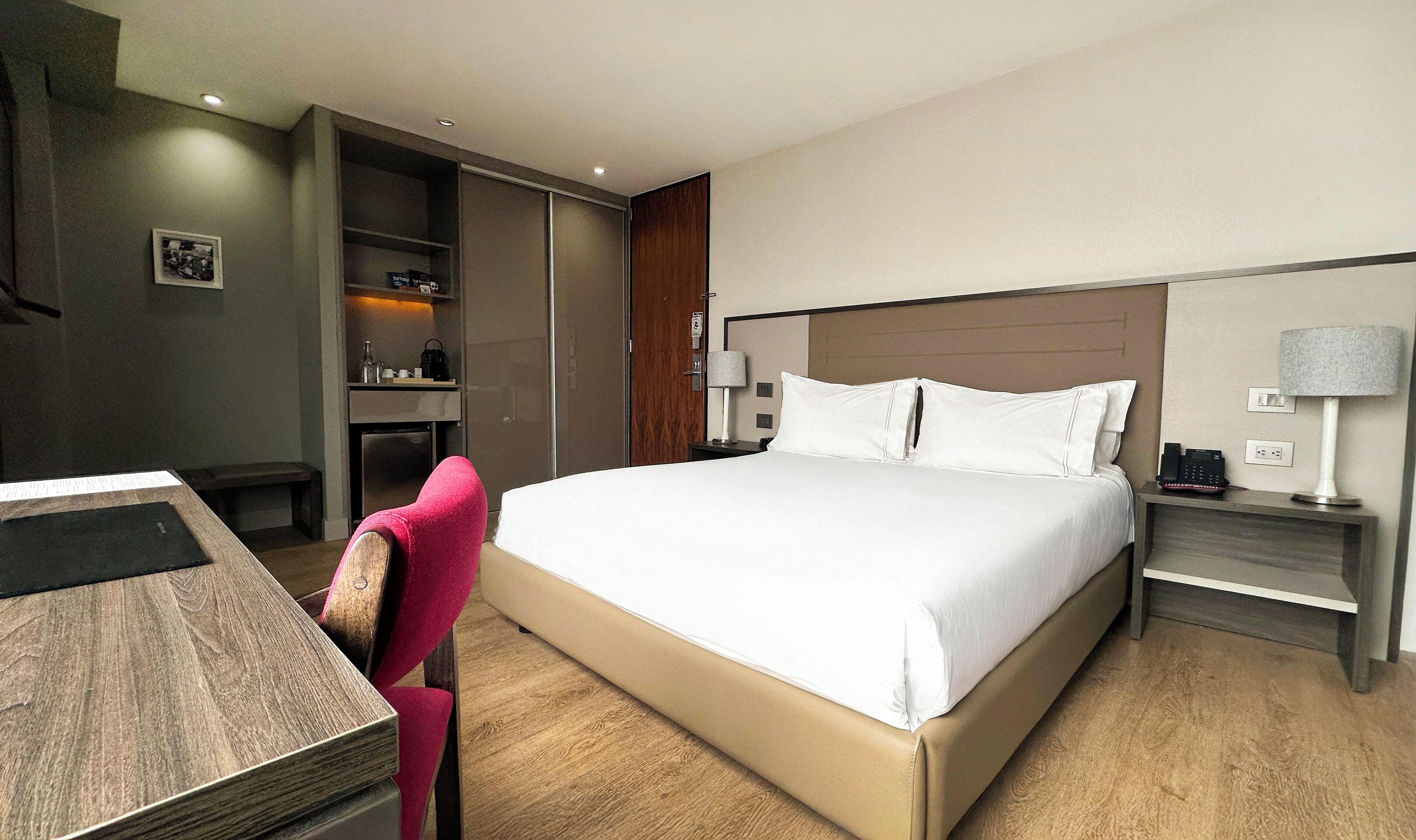 Una habitación de hotel con cama, escritorio, silla, mesita de noche, lámpara y armario.