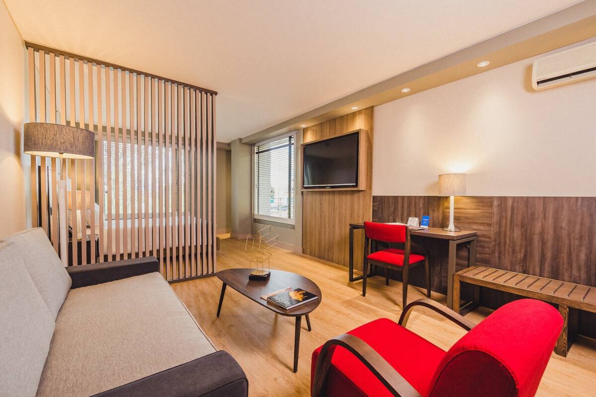 A modern hotel room with a sofa, red chairs, a TV, and a wooden partition.