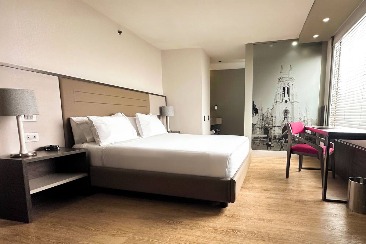 A hotel room with a bed, bedside tables, a desk, a chair, and a large window with blinds.