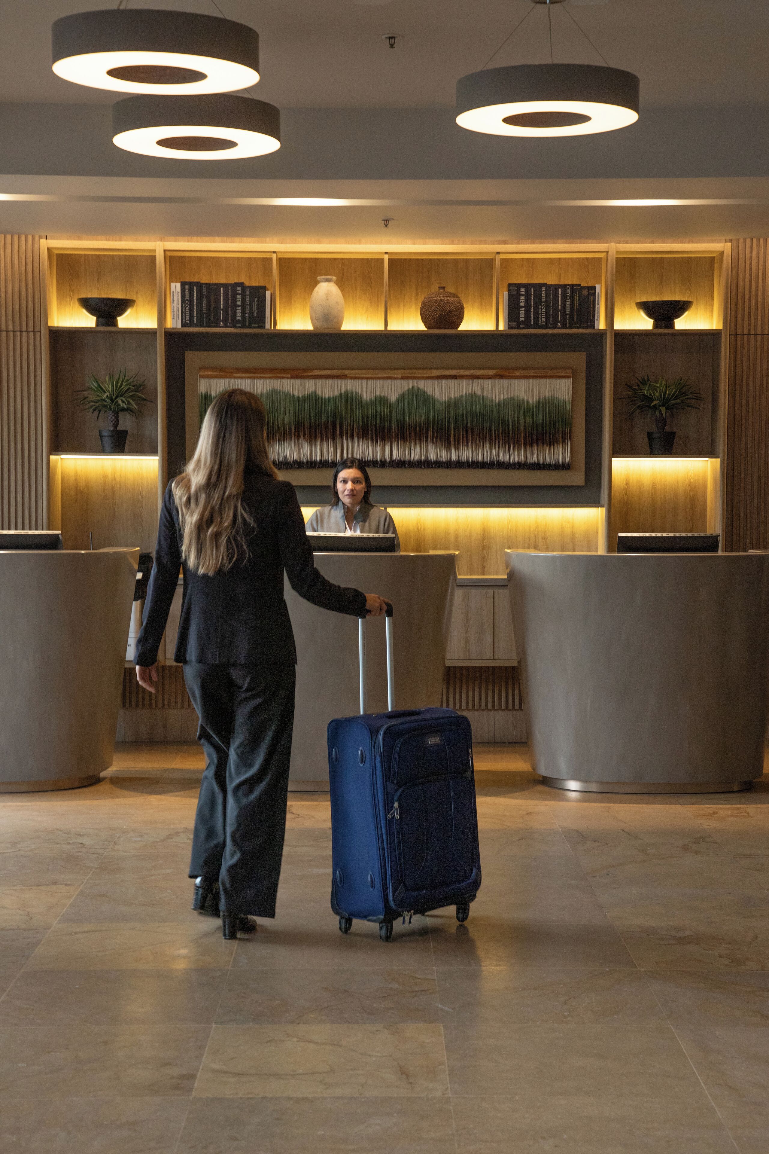 A reception area with a receptionist, a woman with luggage, and a reception desk.