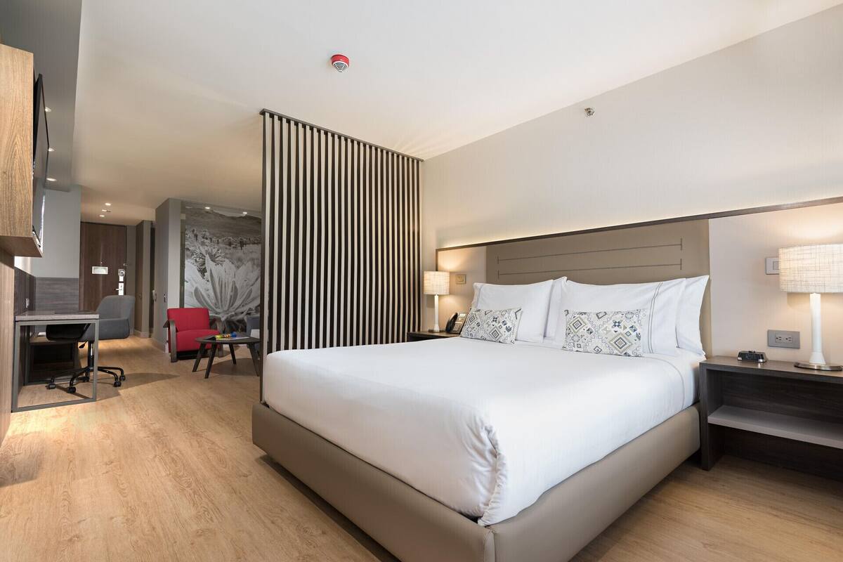 A modern hotel room with a large bed, a desk, a chair, and a red armchair.