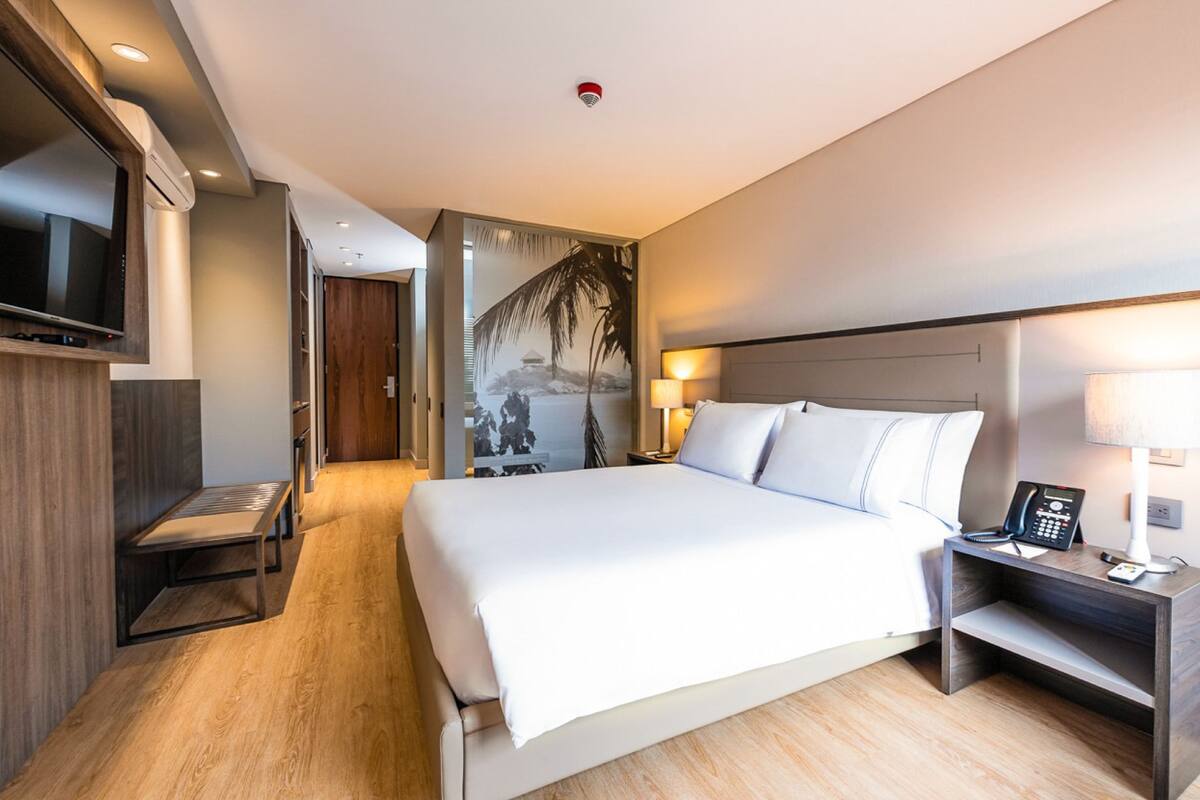 A modern hotel room with a large bed, bedside tables, a TV, and a painting on the wall.