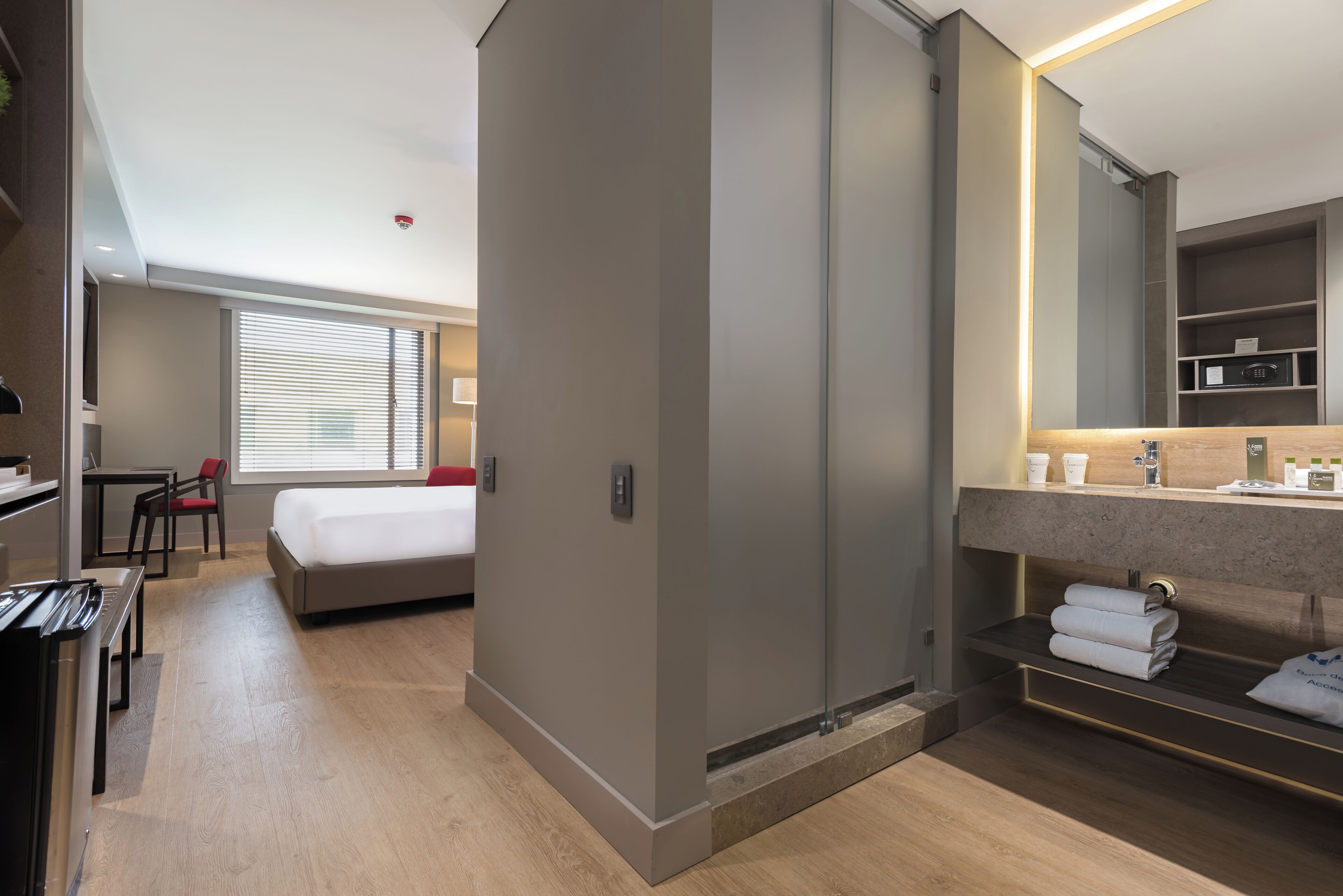 A modern hotel room with a bed, bedside table, and a large mirror. There is a desk with a chair and a window with blinds.