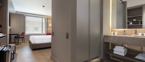 A modern hotel room with a bed, bedside table, and a large mirror. There is a desk with a chair and a window with blinds.