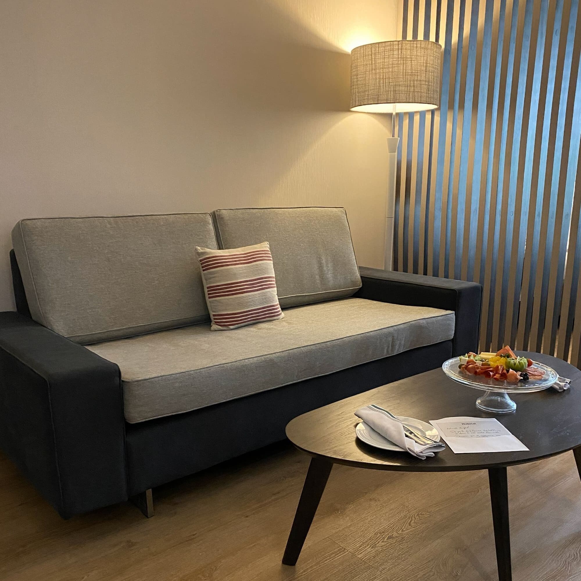 Una sala moderna con un sofá gris, un cojín a rayas, una mesa con un recipiente de vidrio con fruta y una lámpara.