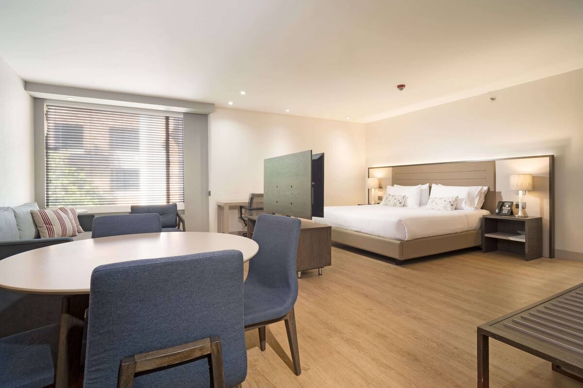 A modern hotel room with a bed, bedside tables, a desk, a dining area with chairs, and a large window with blinds.