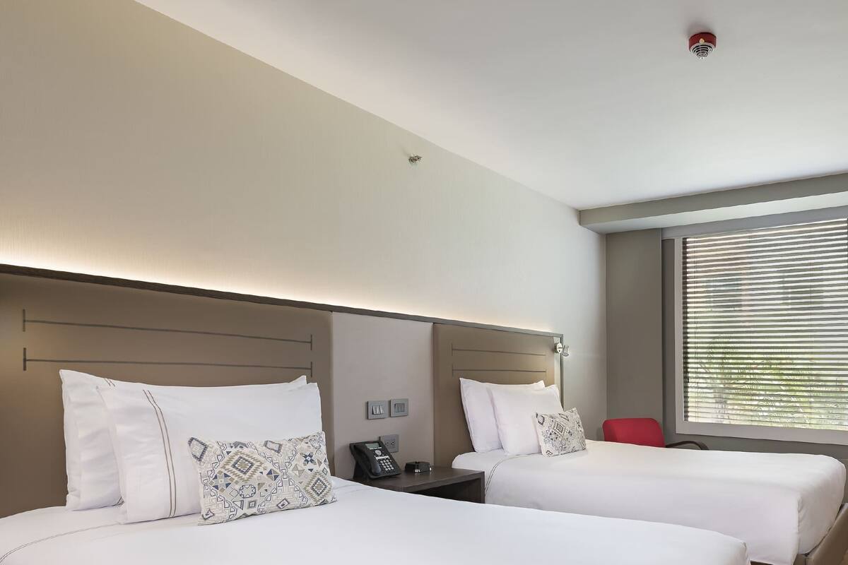 A hotel room with two beds, a bedside table with a phone, and a window with blinds.