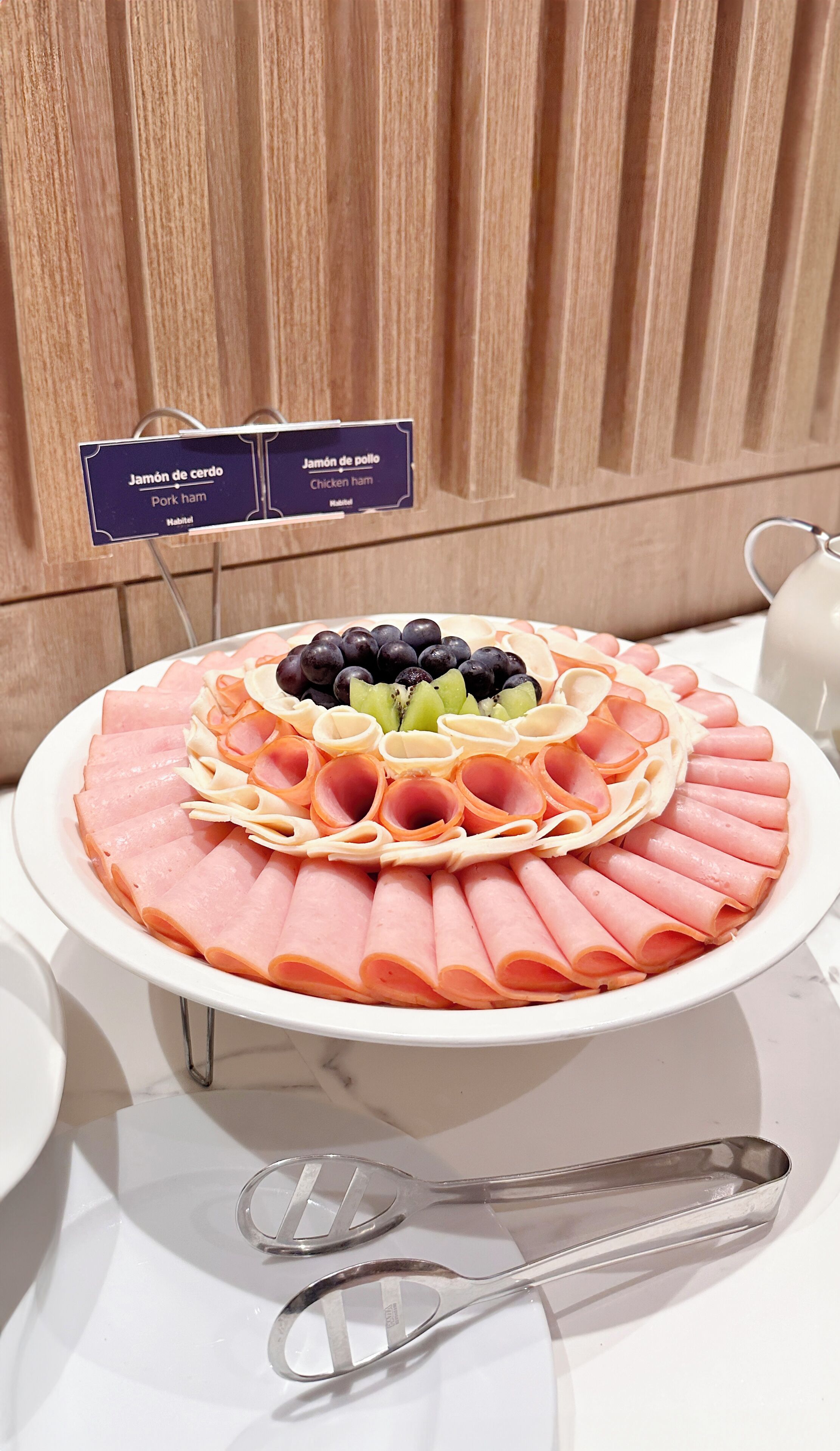 Una bandeja con fiambres y frutas cortadas sobre un plato blanco, con un letrero que indica 'Jamón de cerdo' y 'Jamón de pollo'.