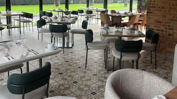 A covered outdoor seating area with tables and chairs, a patterned floor, and a view of a green landscape.