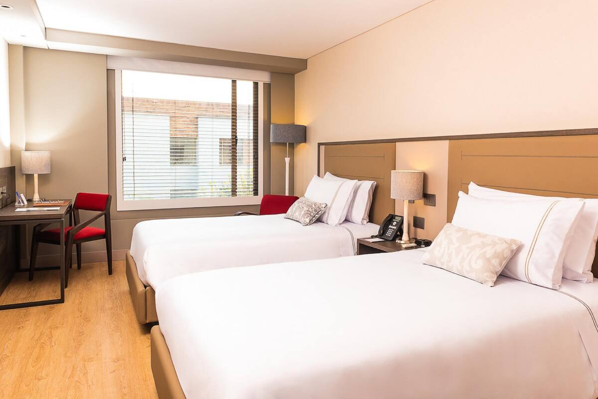 A hotel room with two beds, a desk, a chair, and a window with blinds.