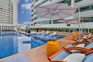 Piscina con sillones de descanso y sombrillas, rodeada por un edificio moderno con grandes ventanales.