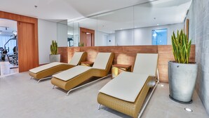 Un área de espera moderna con sillones de mimbre, una planta grande en maceta y un gimnasio visible al fondo.