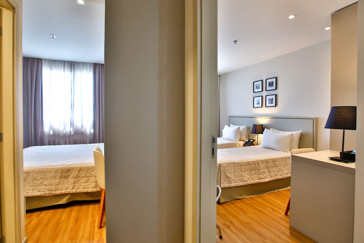 Habitación de hotel con dos camas, una mesita de noche con lámpara, una silla y un ventanal con cortinas.