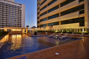 Uma área de piscina de hotel com um elemento aquático, espreguiçadeiras e um edifício com vários andares.