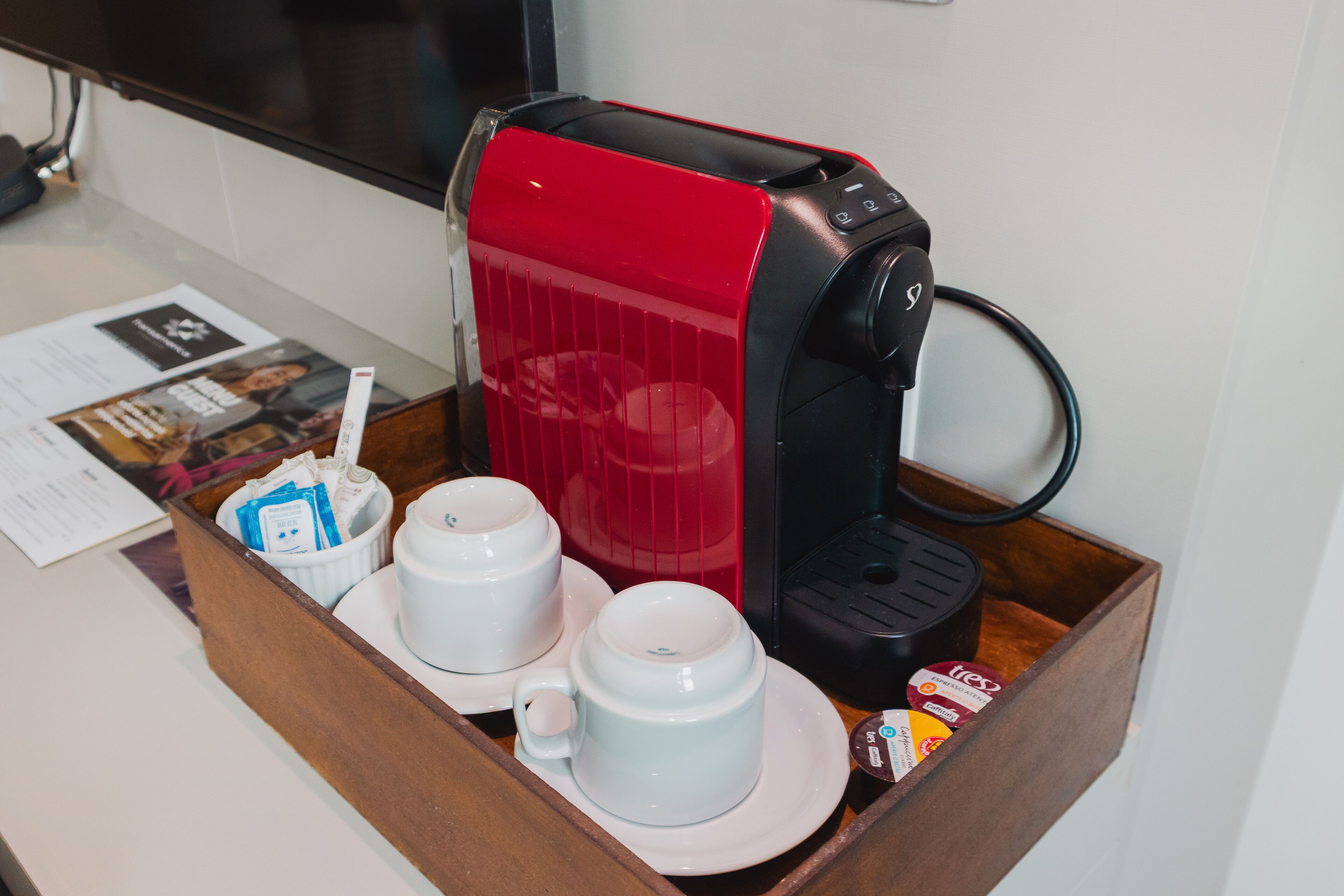 En kaffebryggare med ett rött lock, en kaffekapsel och två vita koppar på ett fat.