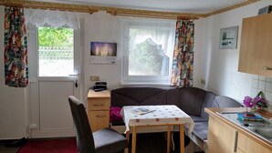 Una habitació petita i acollidora amb un llit individual, una taula amb un mantell de punts, una cadira, un petit armari i una finestra amb cortines.