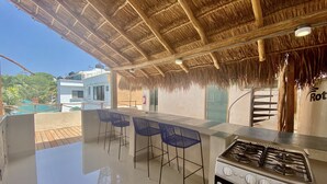Uma cozinha externa com fogão a gás, banquetas de bar e um telhado de palha.