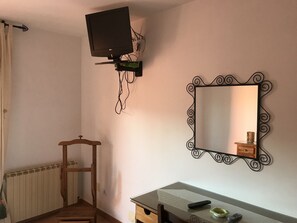 A room with a television mounted on the wall, a mirror with a decorative frame, a wooden chair, a small table with a glass top, and a radiator.