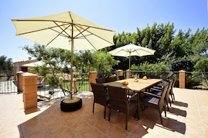 An outdoor patio with a table and chairs, two large umbrellas, and a view of trees and a building in the background.
