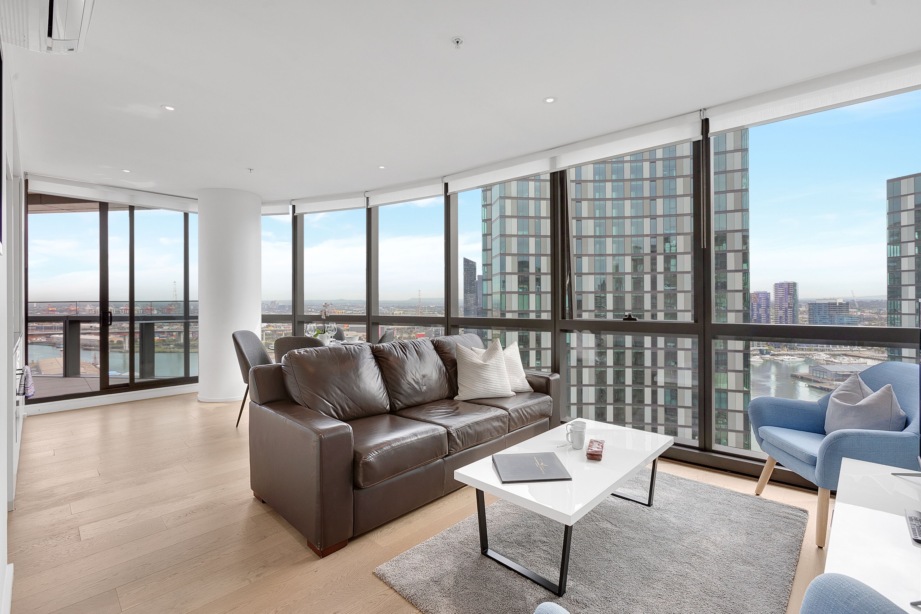 A spacious living room with a brown leather sofa, a white coffee table, and a blue armchair. Large windows offer a view of a cityscape.