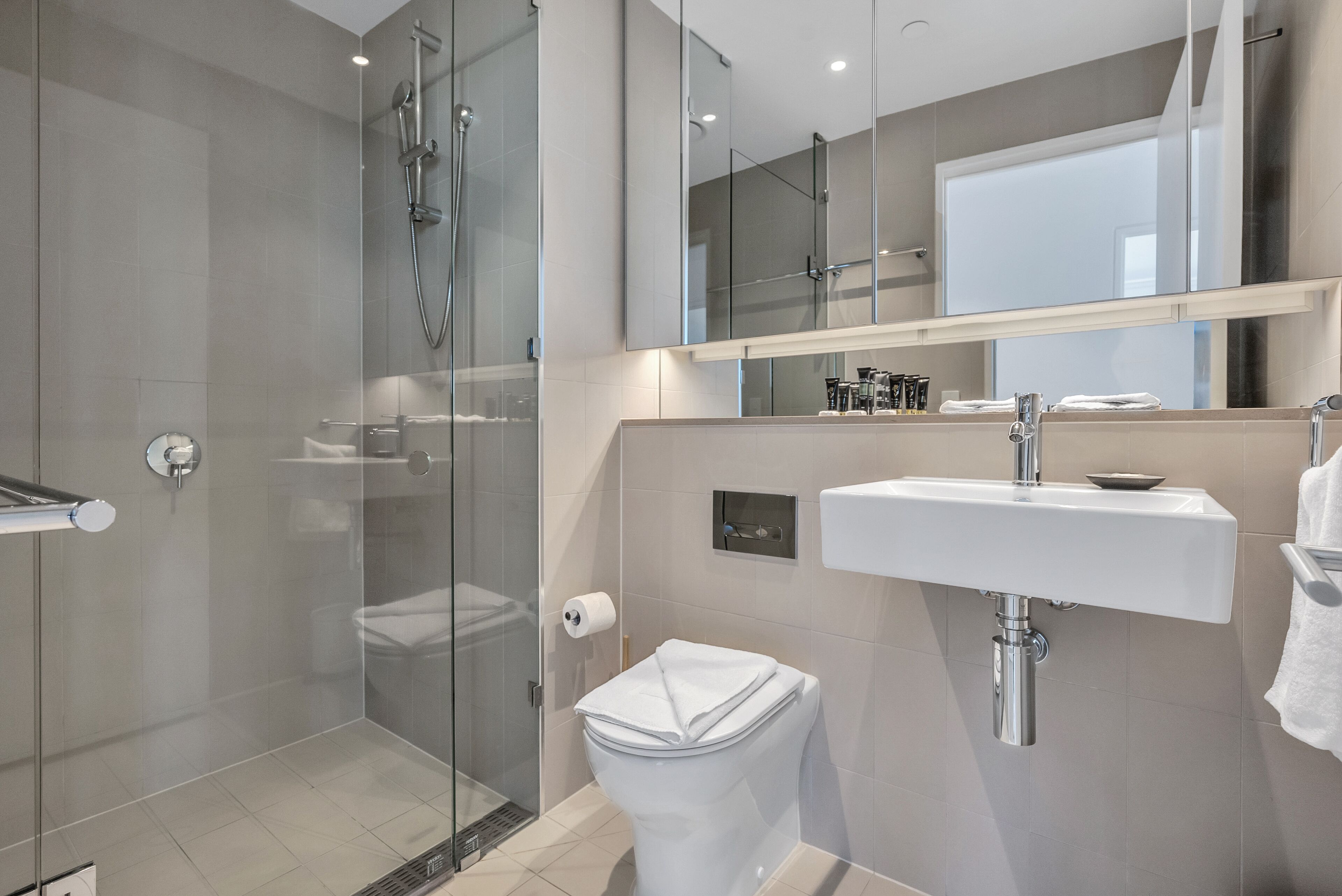 Ein modernes Badezimmer mit einer gläsernen Duschkabine, einer weißen Toilette, einem Waschbecken mit Spiegel darüber und einem Handtuchhalter.