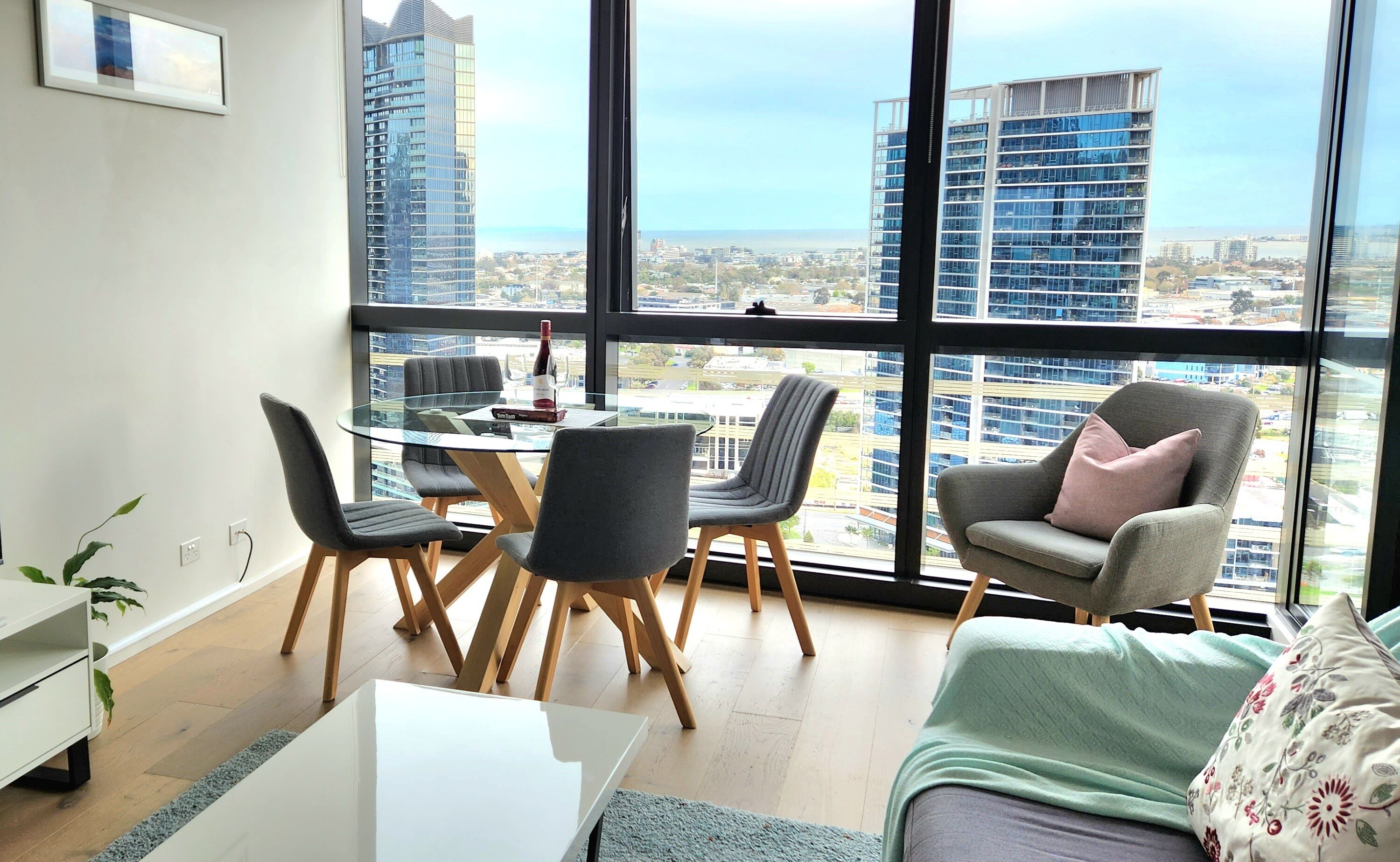 A modern living room with a glass table, four chairs, a sofa, and a view of a cityscape through large windows.