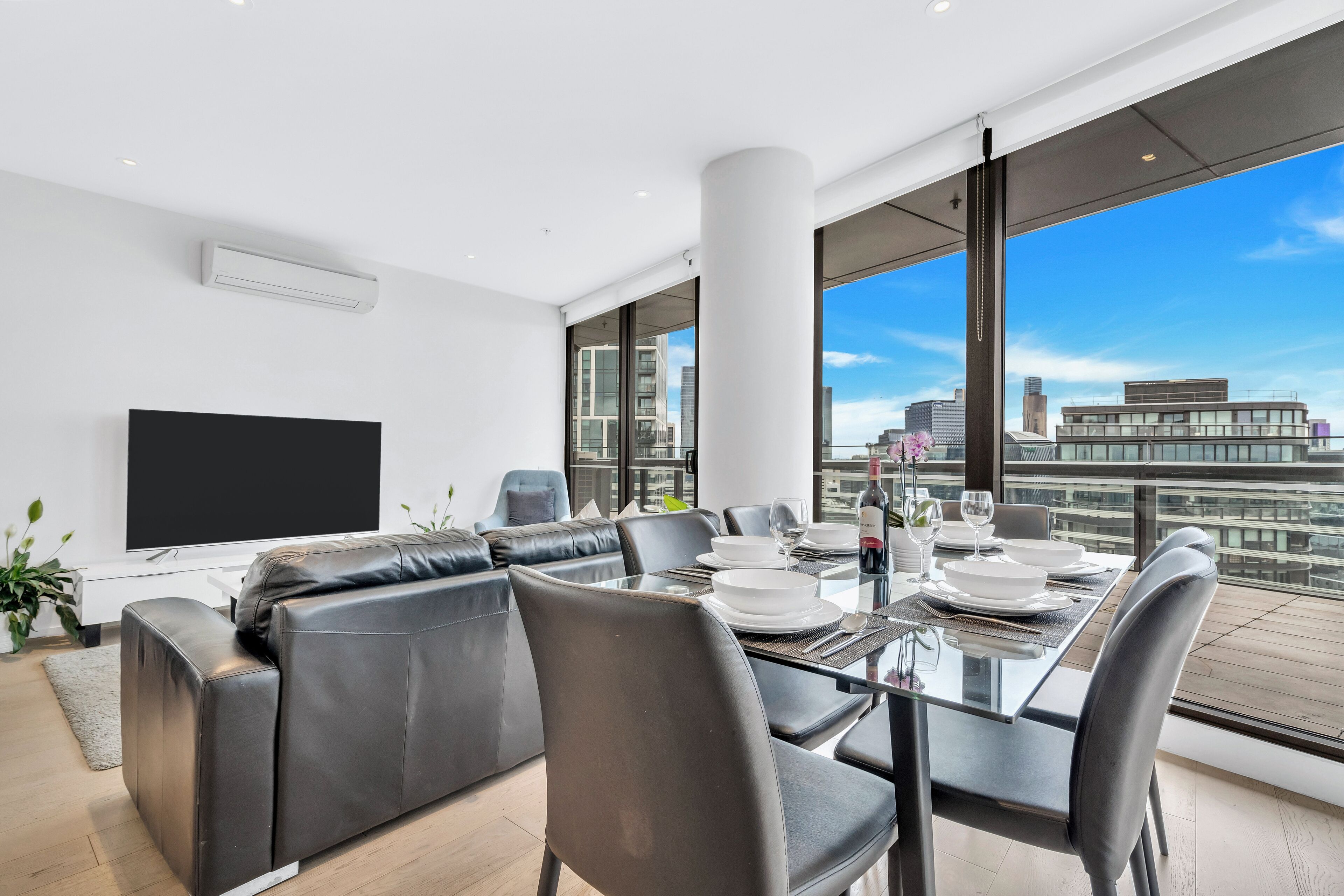 A modern living room with a glass dining table, leather sofa, and a large flat-screen TV.