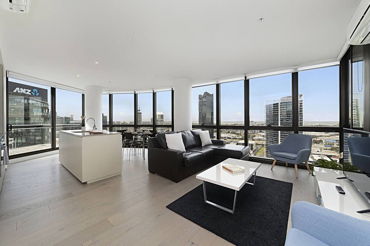 Un salon moderne avec un canapé en cuir noir, une table basse blanche et une vue sur les gratte-ciel de la ville, visible à travers de grandes fenêtres.