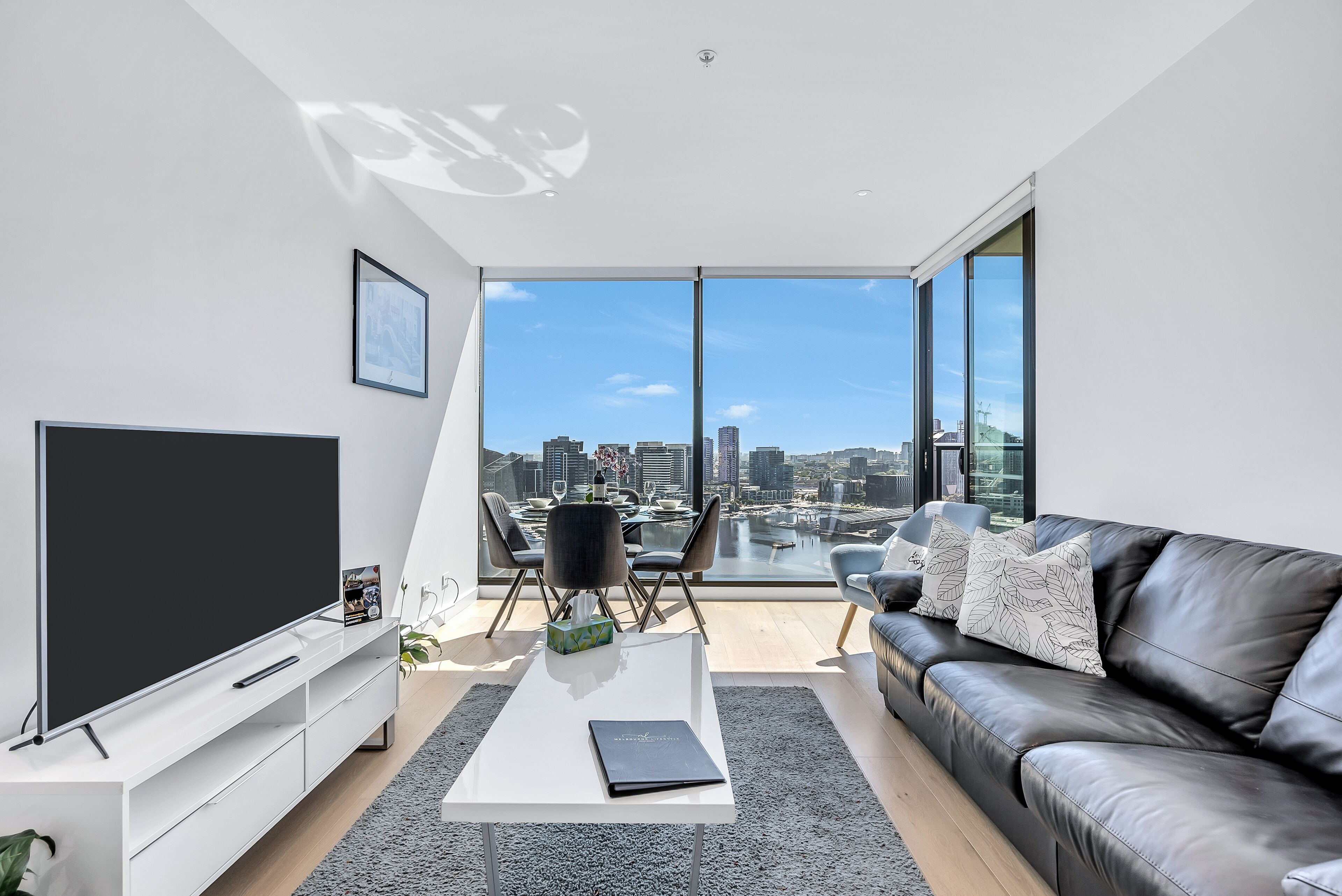 A modern living room with a large TV, a black leather sofa, a white coffee table, and a dining area with a view of the city skyline.