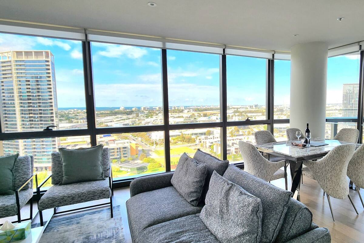 Une chambre d’hôtel moderne dotée de grandes fenêtres donnant sur la ville, d’un coin repas avec une table et des chaises, et d’un canapé agrémenté de coussins.