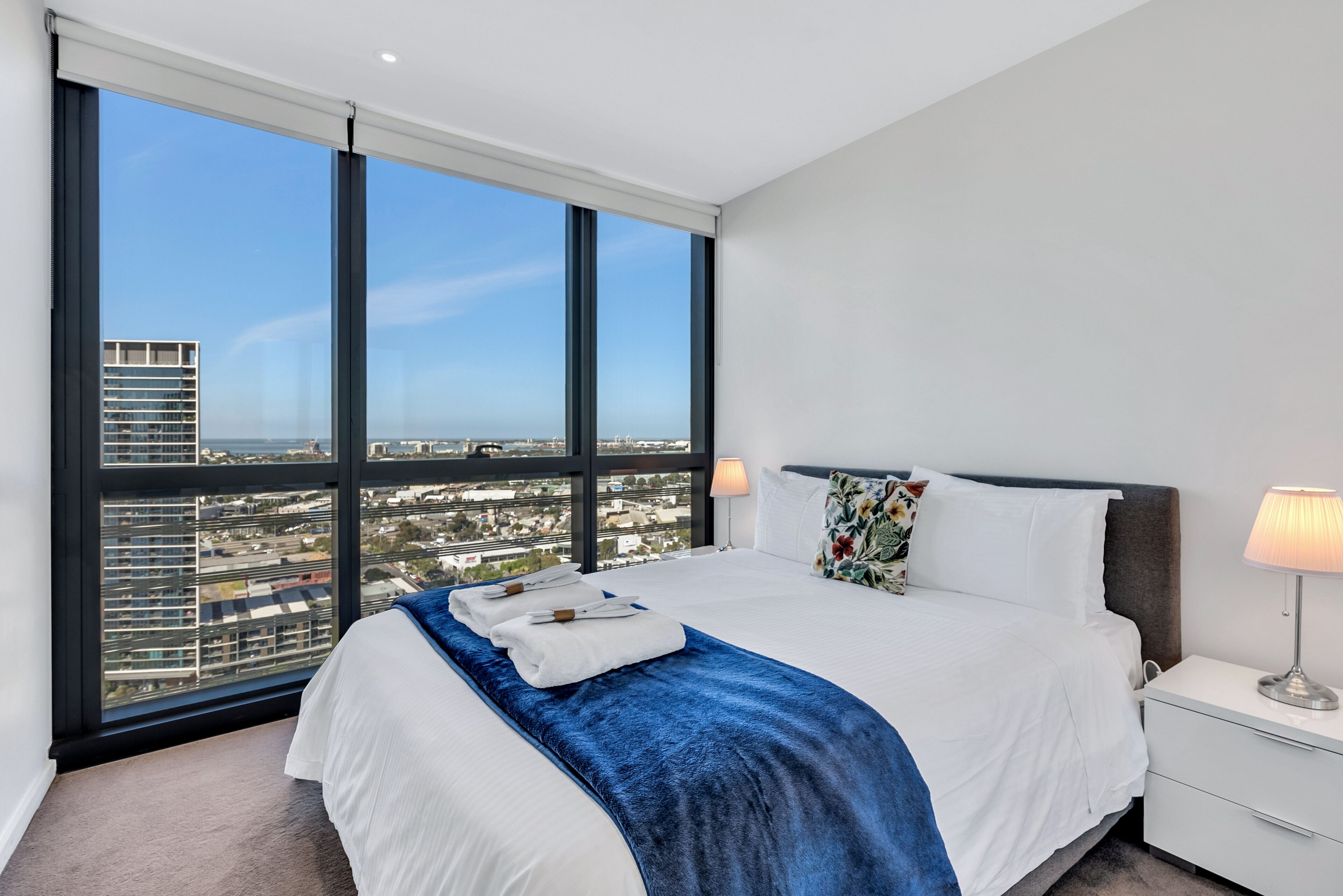 Une chambre d’hôtel moderne dotée d’un grand lit, d’une table de chevet et offrant une vue sur la ville grâce à de grandes fenêtres du sol au plafond.