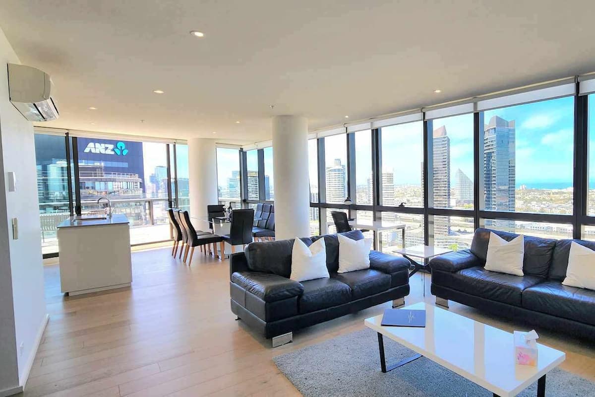 Un salon moderne comprenant un ensemble de canapés, une table basse et un coin repas avec des chaises. De grandes fenêtres offrent une vue sur le paysage urbain et les bâtiments.