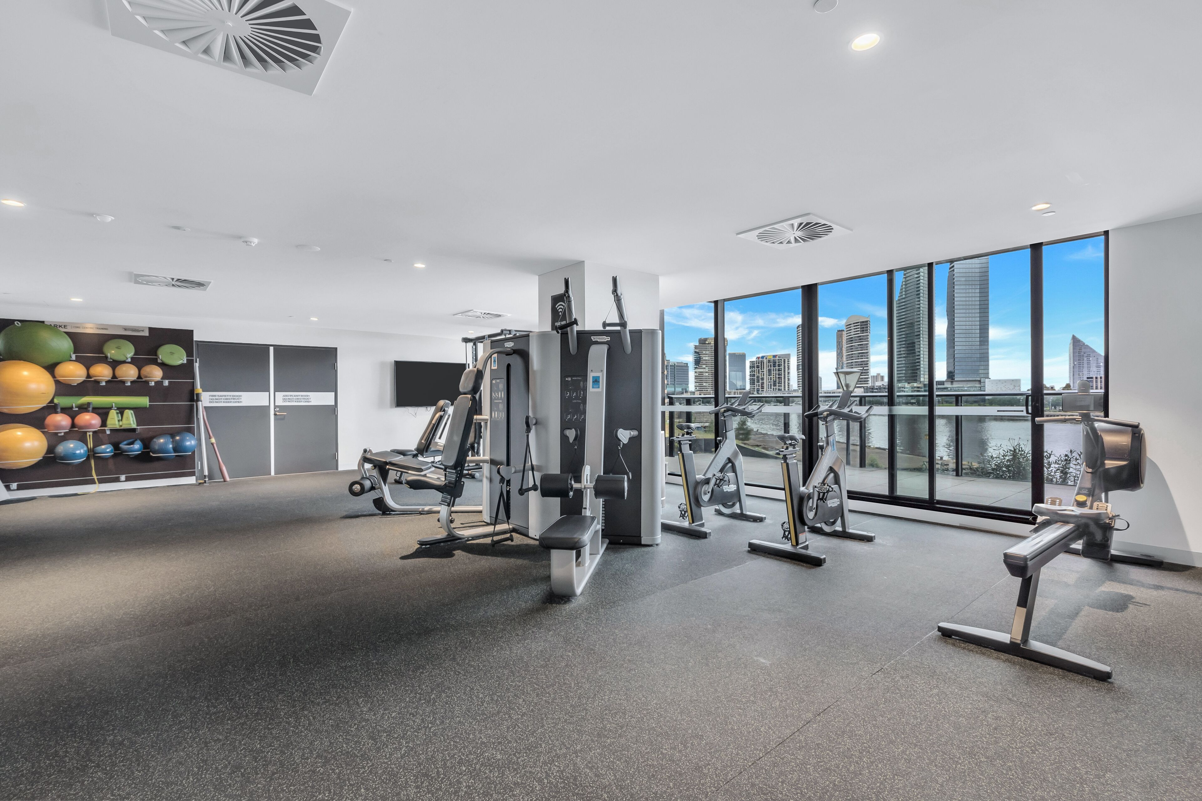 A modern gym with various exercise equipment, large windows offering a city view, and a wall with exercise balls.