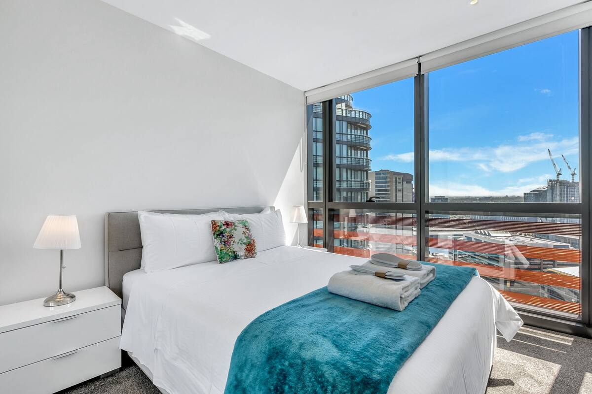 Une chambre d’hôtel moderne avec un grand lit, une table de chevet, une lampe et une vue sur le paysage urbain grâce à de grandes fenêtres.