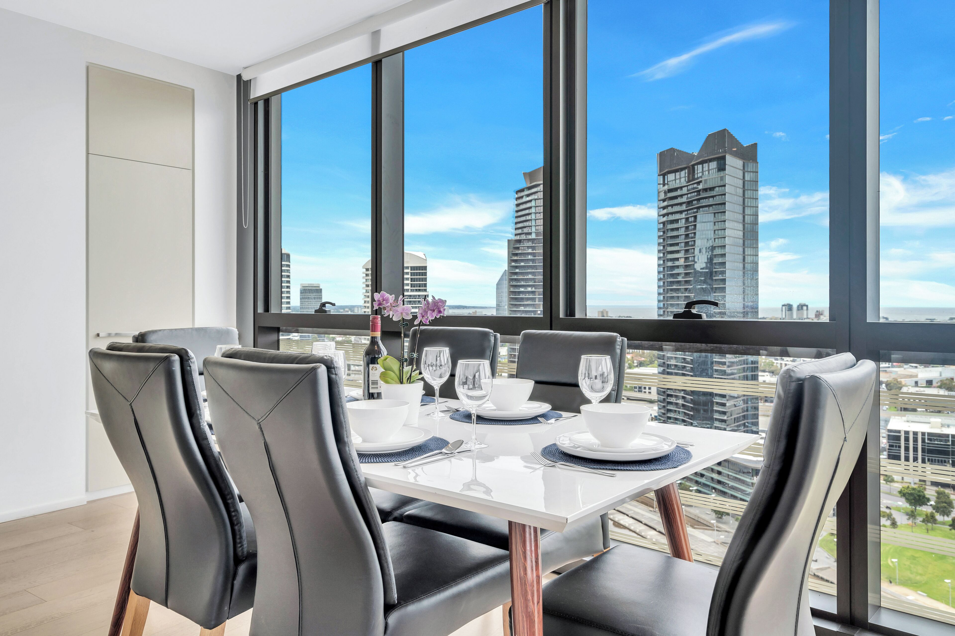 Un coin repas avec une table dressée pour quatre personnes, de grandes fenêtres offrant une vue sur la ville et des chaises noires au design moderne.