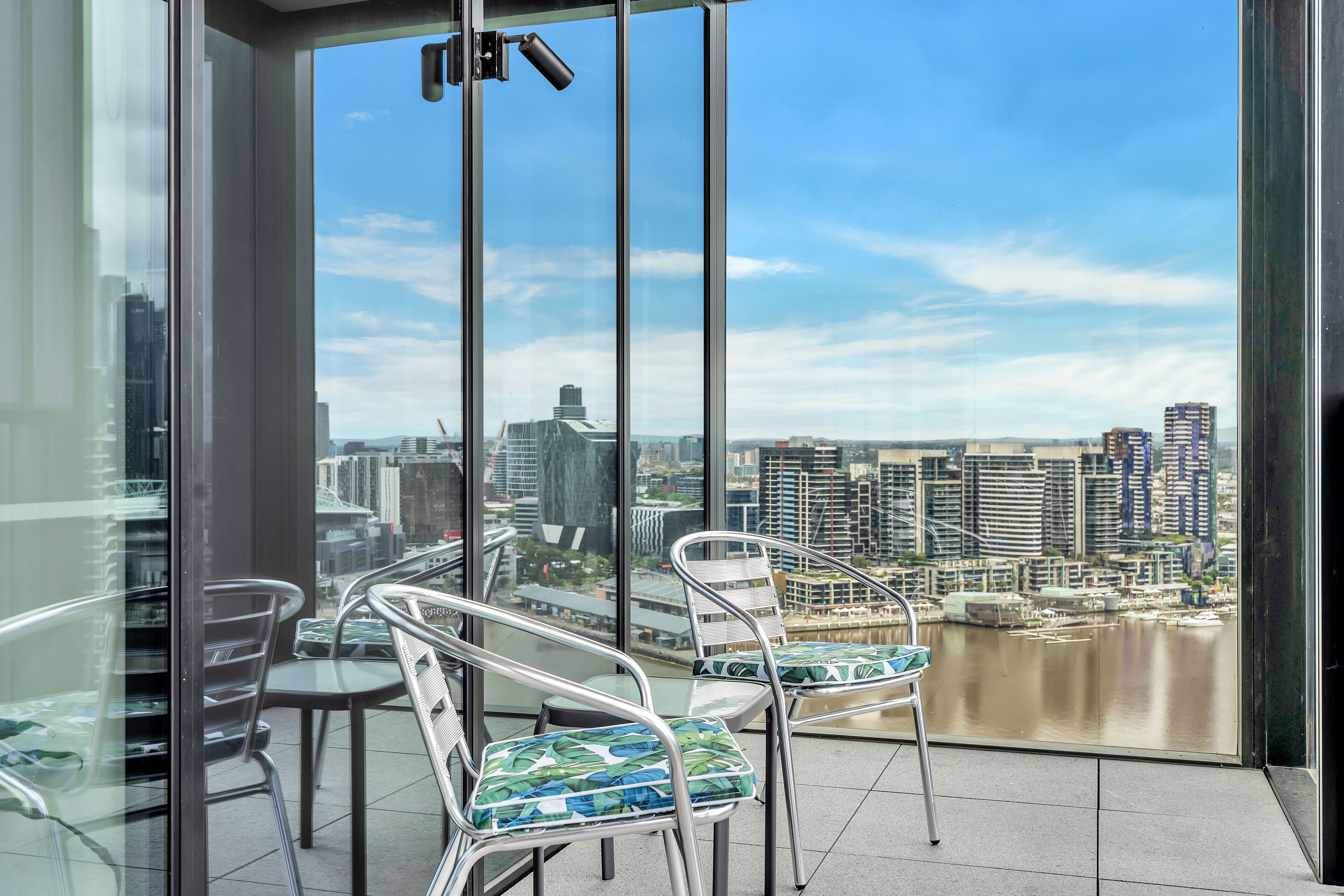 Un balcón con vista a la ciudad, sillas de metal y una mesa de vidrio.