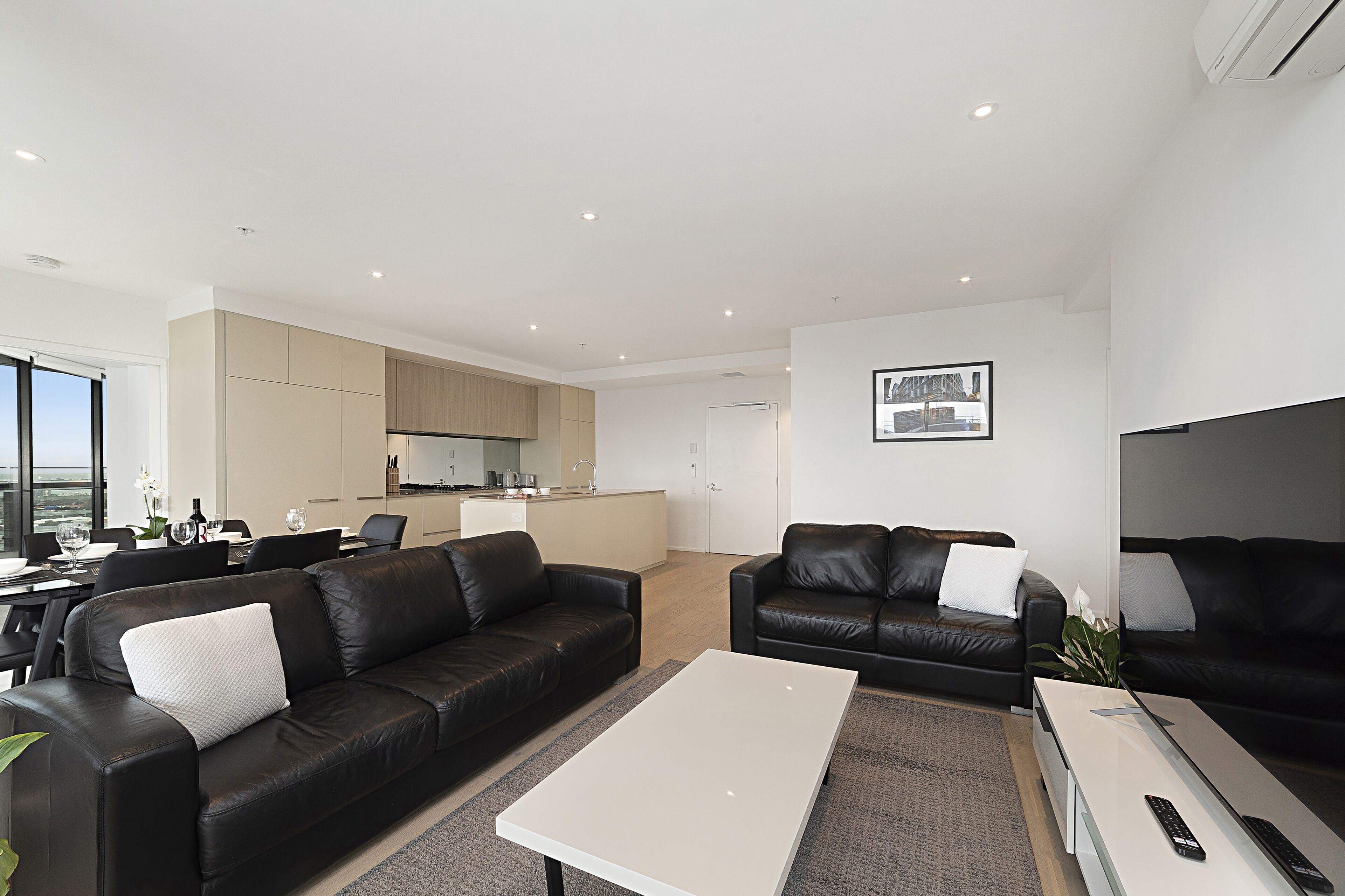 A modern living room with black leather sofas, a white coffee table, and a kitchen area in the background.