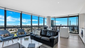 Ein modernes Wohnzimmer mit einem schwarzen Ledersofa, zwei blauen Sesseln, einem weißen Couchtisch und Blick auf die Stadt durch große Fenster.